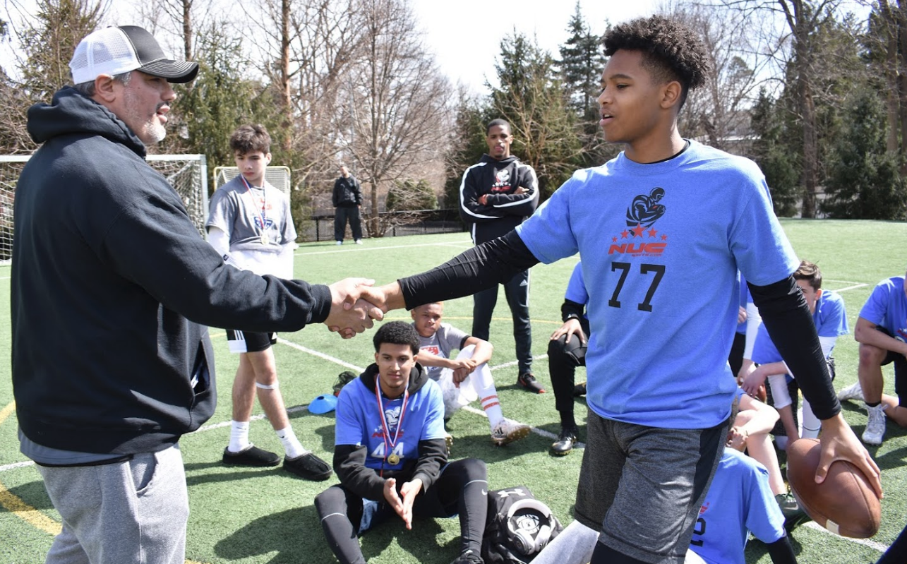 Coach David Schuman, founder of NUC Sports, shaking hands with a high school football prospect in pads and helmet on the sideline of a combine before warm-ups begin.