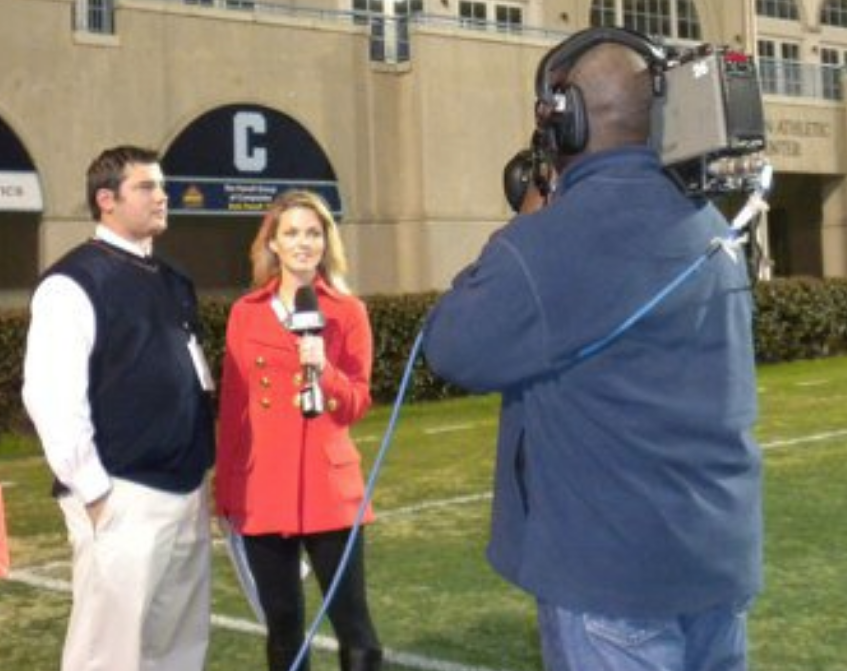 Coach David Schuman speaking into a broadcast microphone during a sideline interview at NUC All-American week, breaking down prospect performance on a live feed.