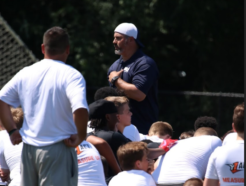 Portrait of Coach David Schuman, founder of NUC Sports and host of The Prospect Podcast, speaking to a room of football prospects in a black NUC polo.